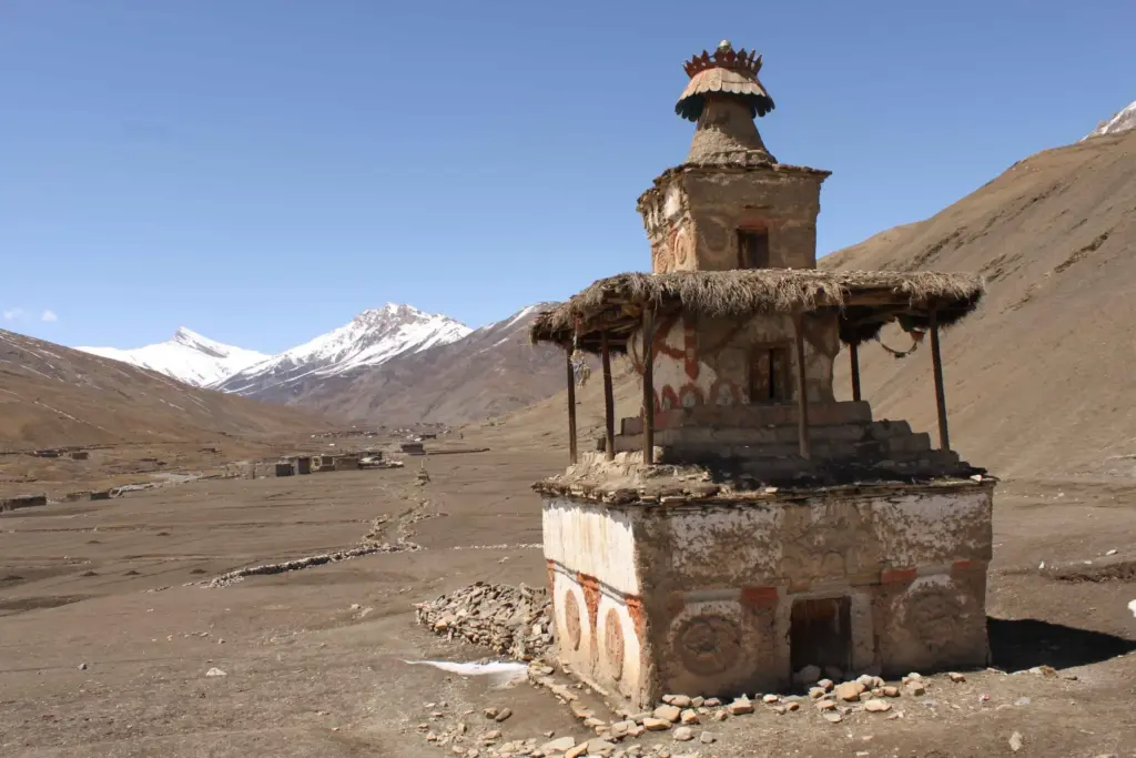 A traditional stone Buddhist chorten stands in a dry, rain-shadow valley landscape with snow-capped mountains in the background, a perfect destination for monsoon treks in Nepal.