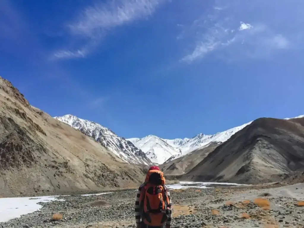 Rear view of a hiker with a large orange backpack trekking through a rocky riverbed valley toward snow-capped mountain peaks under a bright blue sky.
